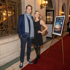 Michael Engelman and Tamara York at the film festival gala. He's in a navy blue blazer, gray scarf, and casual shoes. She wears a black sleeveless dress with stockings, standing next to an easel.