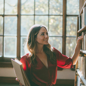 Claire Forlani, wearing a red silk blouse, standing beside a bookshelf in a cozy library. She has a kind smile and long wavy hair. Background features large windows with sunlight filtering through.