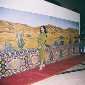 Teresa Ruiz in green dress and beige heels at the 'Narcos: Mexico' premiere. Desert-themed artwork backdrop, red carpet event.