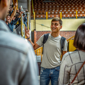 Paulo Dybala at an autograph signing event. Wearing a casual outfit, jeans and t-shirt. The backdrop is filled with cheering fans.