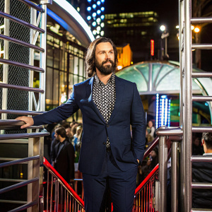 Milo Ventimiglia, with a beard and long hair, at a press event. Wearing a navy suit and a patterned shirt. Confident pose. Event stage background.