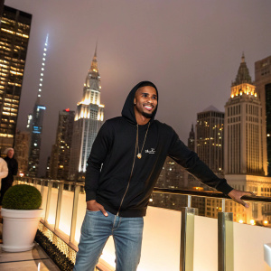 Trey Songz, wearing a black hoodie and jeans, at an upscale gallery opening. Lavish cityscape backdrop