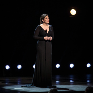 Mayim Bialik, in a stunning black dress, center stage at a gala. Dark background with soft lighting and dimmed spotlights. Microphone at her feet