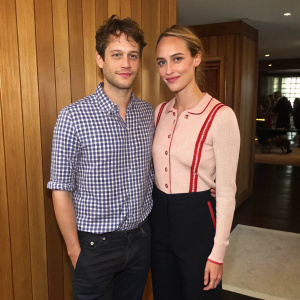 Jonathan Belolo and Nora Arnezeder, close-up candid photo. Jonathan in a checked shirt, Nora in a pastel top with red accents and black trousers. Indoor setting with wooden paneling