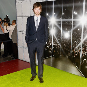 Freddie Highmore at a glitzy awards show, dressed in a sophisticated navy suit with a silver tie. Large crowd and flashing lights in the background.