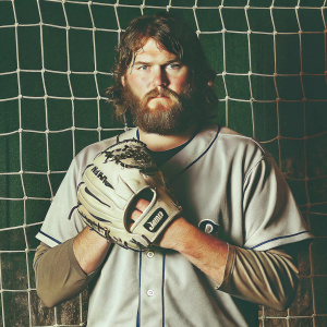 Jackson Kowar, a baseball player with a beard, in a serious pose. Wearing a Kansas City Royals jersey and holding a baseball glove. Dark net backdrop, uniform details, baseball glove details.