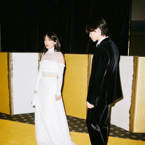 Lee So-Yeon and Nam Gung-Min at a glitzy award ceremony. So-Yeon in a white gown with sheer panels, structured silhouette. Gung-Min in a black velvet blazer. White backdrop with gold accents