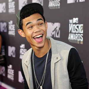 Roshon Fegan, young man with a playful grin, making a funny face at the MTV Video Music Awards. He's dressed casually with a logo-filled backdrop behind him.
