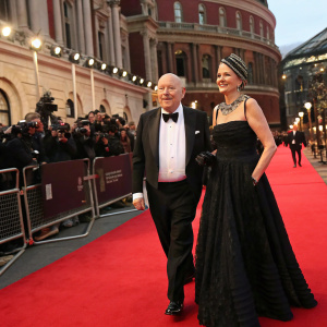 Julian Fellowes and Emma Fellowes attending the BAFTA Film Awards. Julian in a tuxedo, Emma in an elegant black gown with a striking headpiece. The venue is the Royal Albert Hall, with red carpet and flashing cameras.