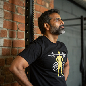South Asian man with beard and serious expression. Wearing black t-shirt with fitness graphic. Brick wall backdrop. Gym setting.