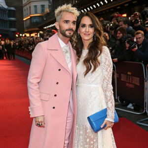 Bill Kaulitz and Johanna Klum at a glitzy Hollywood premiere. Pink coat, white lace dress with blue clutch. Crowded red carpet with paparazzi in the background.