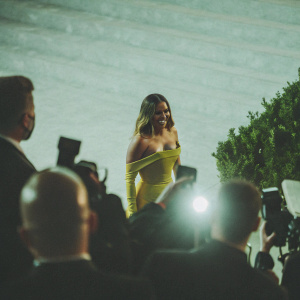 Miley Cyrus at the Met Gala. Wearing a bright yellow, off-the-shoulder dress. Confident stance on the red carpet, surrounded by flashing cameras.