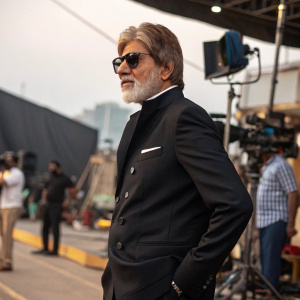 Amitabh Bachchan, distinguished Indian actor, sporting a beard, in a formal black suit and sunglasses. Posing confidently against a bustling film set backdrop, amidst film equipment.