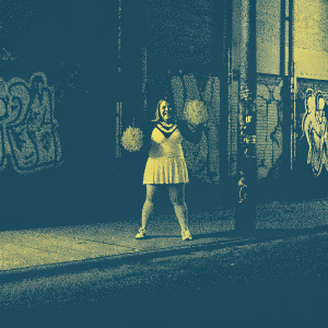 Brec Bassinger, a bubbly actress, with a lively smile and a cheerleader-style outfit. Poses in front of a graffiti-covered wall. The scene is set in an urban alleyway.
