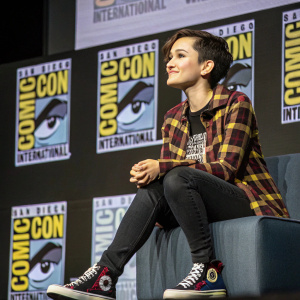 Bex Taylor-Klaus at San Diego Comic-Con. Wearing plaid shirt, dark pants, and sneakers. Prominent Fede Pop and Comic-Con branding visible in background.