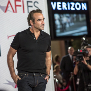 Jean Dujardin, actor, serious expression, in black polo shirt, dark jeans. At a press conference, standing in front of a large digital screen displaying AFI awards and Verizon logos