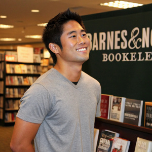 Ryan Higa, wearing a casual gray t-shirt, smiles confidently in a bookstore setting. Shelves of books and the Barnes & Noble logo in the background.