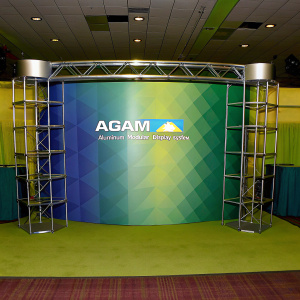 Trade show exhibit featuring AGAM Aluminum Modular Display System. Sleek and modern design with geometric black backdrop. AGAM logo prominently displayed with additional text. Clean lines and simple industrial aesthetic.