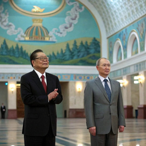 Formal photo of Jiang Zemin and Vladimir Putin at Moscow State University. Jiang Zemin, East Asian male, in dark suit and red tie, glasses on. Putin, Caucasian male, in gray suit and patterned tie. Standing, clapping, in grand hall with intricate blue, green, and gold mural.