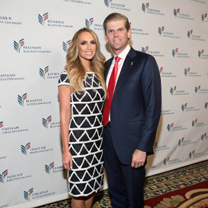 Eric Trump and Lara Yunaska at a charity gala. Eric in a navy suit and red tie, Lara in a black and white geometric dress. They stand against a white backdrop with repeated logos. Patterned floor visible in background.