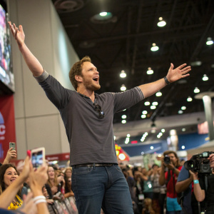 Chris Pratt, arms upraised, at a bustling convention. Casual wear. Excited fans in the background.