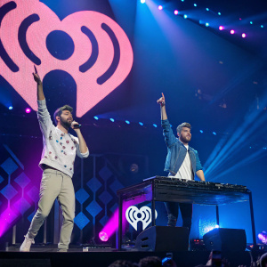 The Chainsmokers, Andrew Taggart and Alex Pall, performing at the Jingle Ball concert. Madison Square Garden stage, illuminated with vibrant blue and pink lights. They are energetically engaged, making dynamic poses with raised arms. Large sound wave design backdrop.