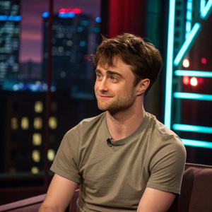 Daniel Radcliffe at an indoor interview, wearing a casual t-shirt. Background features neon city lights