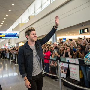 Niall Horan at JFK Airport, waving and smiling at a diverse crowd of fans. Fans of all ages and genders hold up cameras and signs.
