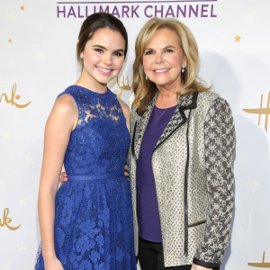 Bailee Madison and Patricia Riley gracing the red carpet at a glamorous Hallmark Channel premiere. Bailee in a striking blue dress with lace overlay, Patricia in a chic patterned jacket. White backdrop with Hallmark Channel logos and crowns adds to the festive mood.