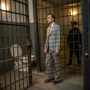 Sheldon Cooper in a plaid suit, standing in a dimly lit jail cell. A police officer is present. The scene feels claustrophobic with large metal bars and a rusted metal toilet.