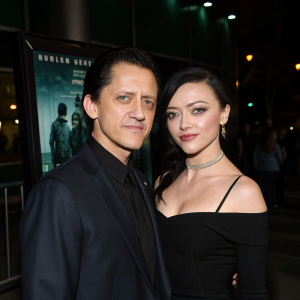 Clifton Collins Jr. and Francesca Eastwood at the Man Down film premiere. Wearing a dark suit and an off-the-shoulder black top respectively. Dark film poster and dimly lit venue in the background. High-quality photo.