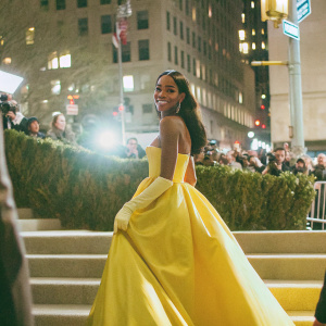 Laura Harrier, at the Met Gala, posing confidently in a vibrant yellow gown. The backdrop is a bustling New York City street at night