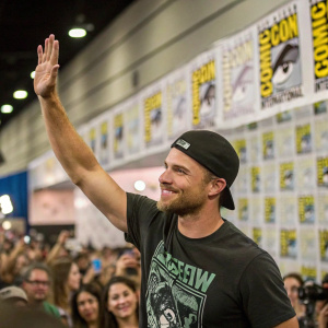 Stephen Amell at a packed Comic-Con booth. Wearing a black cap and graphic t-shirt. He waves enthusiastically. Comic-Con logos plastered on the background.