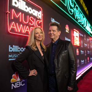 Lisa Kudrow and Chris Isaak at the Billboard Music Awards, posing against a neon-lit backdrop with sponsor logos.