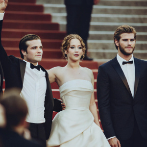 Josh Hutcherson, Jennifer Lawrence, and Liam Hemsworth at a glamorous awards show. They stand in a row, dressed in formal attire, with red carpet stretching out behind them.