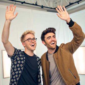 Tyler Oakley and Alfie Deyes, both smiling and waving. Backdrop is a bright studio setting.