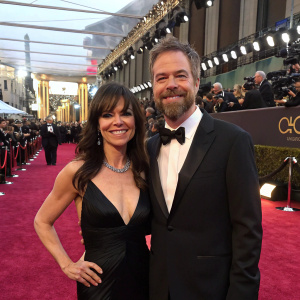 Liz Vassey and David Emmerichs at the Oscars, dressed in black tie and gowns, smiling broadly on the red carpet.