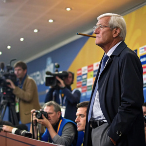 Marcello Lippi, serious expression, holding a cigar-like object. Press conference during World Cup event.