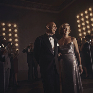 Dominic Chianese and Edie Falco at the award ceremony. They are dressed in formal attire. The venue is illuminated with golden lights, creating a warm and glamorous ambiance.