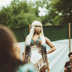 Nicki Minaj at the MTV Video Music Awards, styled in a bold, multi-hued outfit with fringe and tassels. Standing confidently with a direct gaze, platinum blonde wig, against a simple black backdrop.