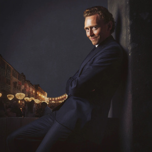 Tom Hiddleston, in a sleek navy suit, posing against a bustling city street background at an outdoor festival.