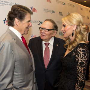 Formal event photo of Rick Perry, Sheldon Adelson, and Miriam Ochsorn. Rick in gray suit, red tie, Sheldon in dark suit, brown hair, Miriam in black detailed gown, blonde hair. They are engaged in conversation. The backdrop features corporate logos.