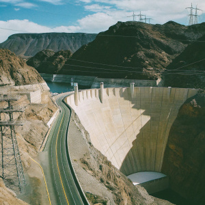 Imposing Hoover Dam standing tall amidst the rocky terrain. The winding road leading up contrasts against the vast blue sky and scattered clouds. The dam's massive structure dominates the scene, surrounded by towering mountains with their rugged, earth-toned peaks.