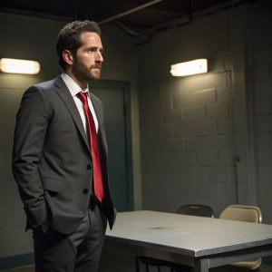 Aaron Abrams as Matthew Weitz in Blindspot. Wearing a dark gray suit and red tie, standing in a dimly lit interrogation room.