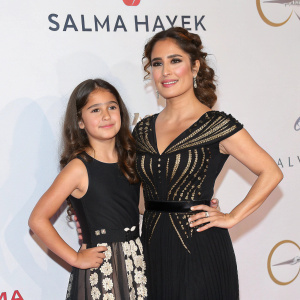 Salma Hayek, in a black dress with intricate details, and a young girl in a black belt-accented dress, pose for the camera at a glamorous event. The backdrop features logos of several sponsors against a clean white surface.