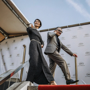Katherine Waterston and Danny Boyle at a film festival. Katherine in a black polka dot dress, short hair. Danny in a suit with glasses. They pose against a white backdrop with event logos.