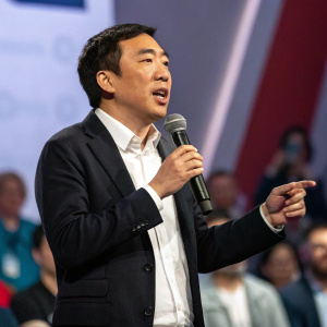Andrew Yang, Asian male, speaking passionately into microphone at tech conference. Dark suit, white shirt, blurred audience in background.
