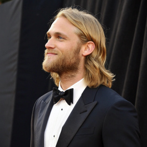 Wyatt Russell, fair-skinned actor with long blonde hair and beard. Formal wear, standing against black backdrop at award show.