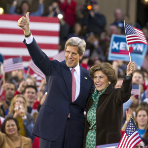 Formal photo of John Kerry and Teresa Heinz-Kerry giving a thumbs-up sign at a rally. Diverse crowd, waving American flags.