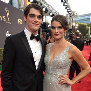 RJ Mitte and Dyna Mitte on the Emmy Awards red carpet. RJ in a black tuxedo and bow-tie, Dyna in a stunning silver sequined gown. The backdrop is lit with cameras and flashing lights.